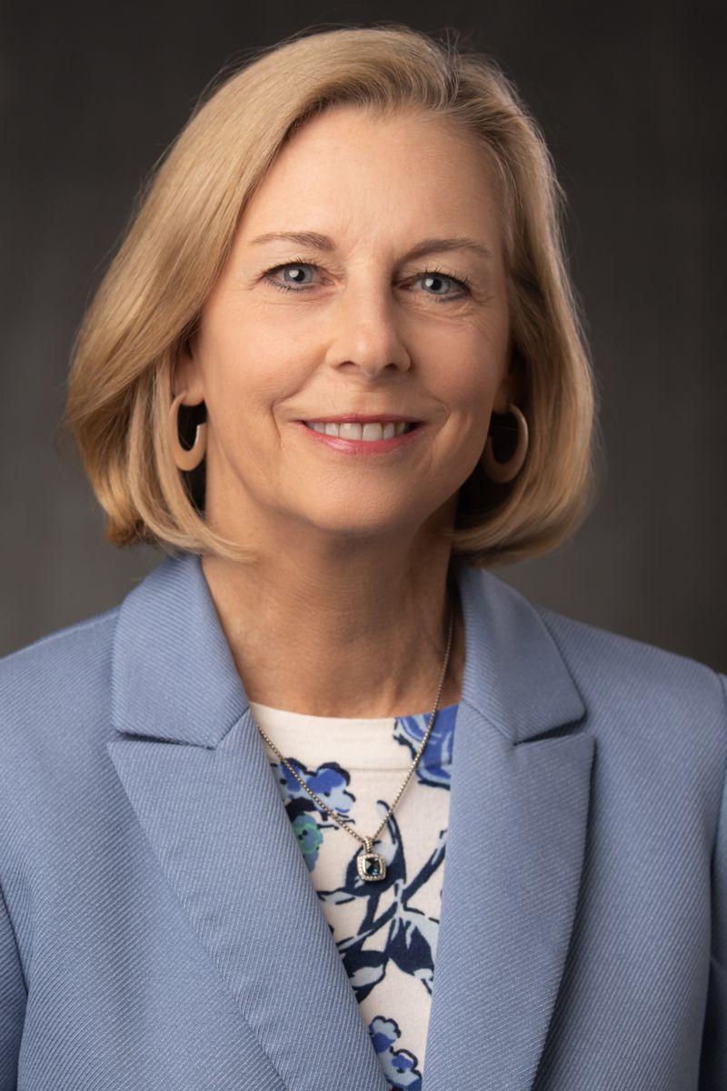 Tammy Shepherd, 戴着金耳环的金发短发白人女性,a silver necklace with a blue stone, a white shirt with blue patterns, and a blue suit jacket.